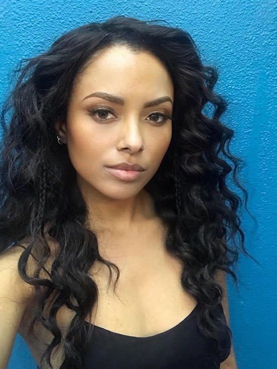 a woman with long black curly hair standing in front of a blue wall and looking at the camera