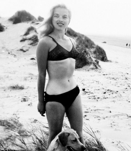 a woman in a bathing suit standing next to a dog on the beach with sand dunes behind her