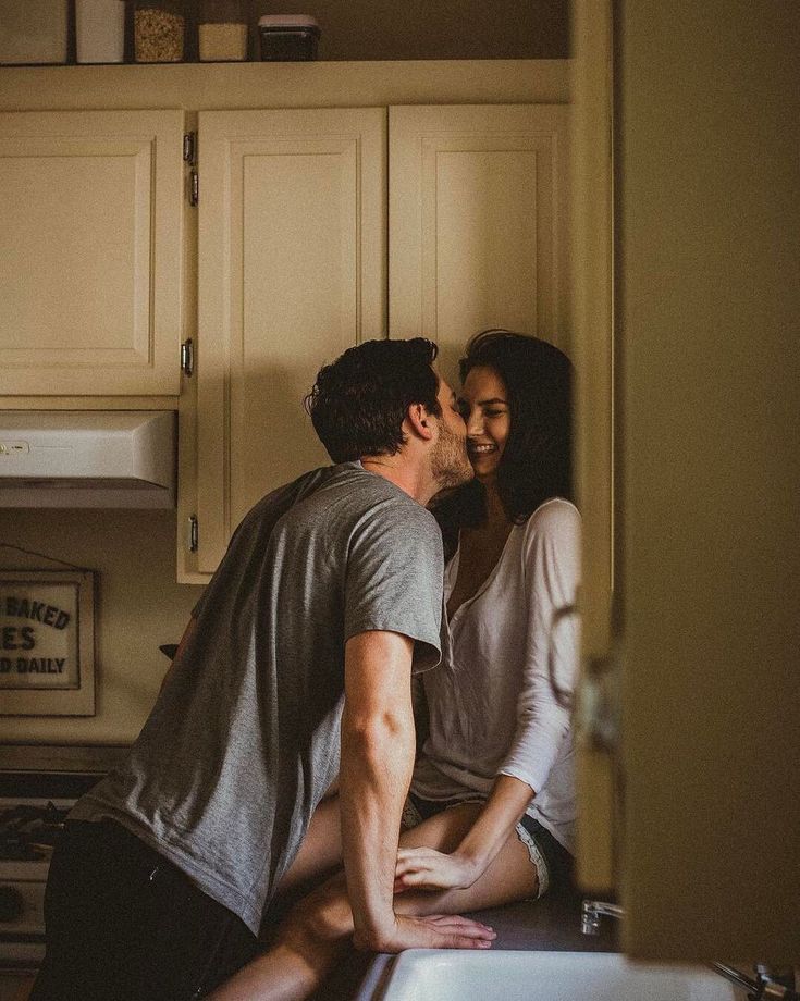 a man and woman kissing in the kitchen while sitting next to each other on the counter