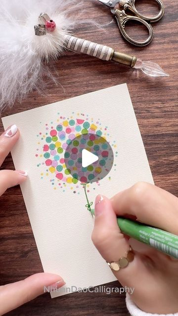 someone is making a card with dots and a balloon on the paper, while another person holds a green marker