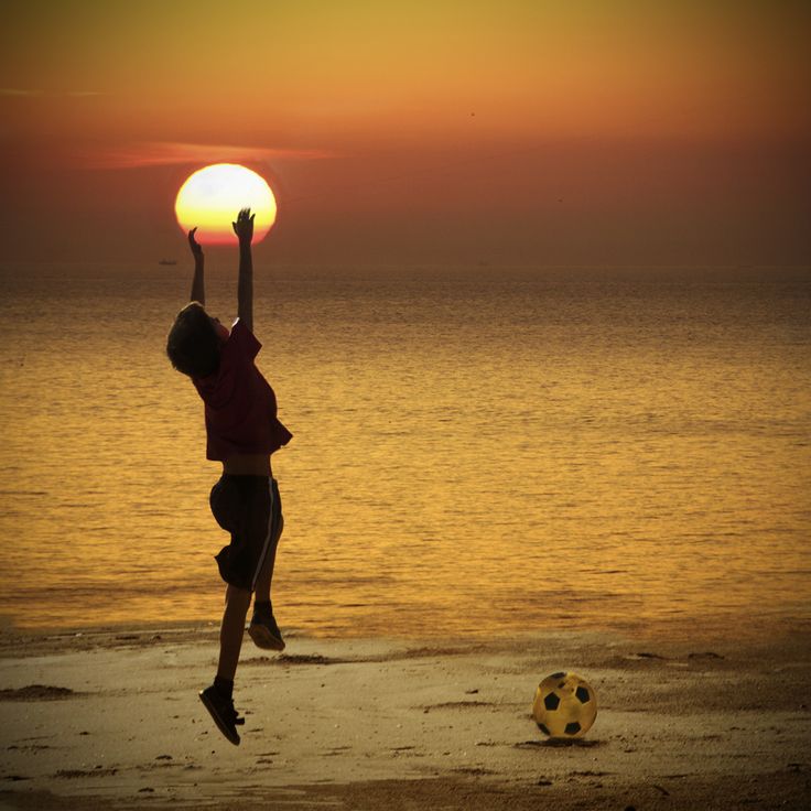 a person jumping up in the air with a soccer ball next to them on a beach