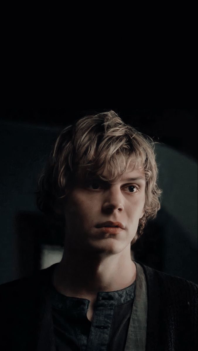 a young man with blonde hair wearing a black shirt and vest looking at the camera