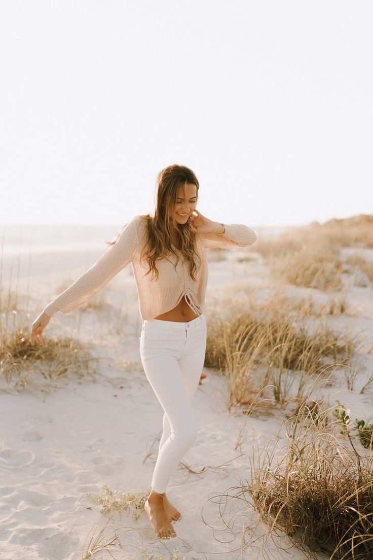 a woman standing in the sand with her arms behind her head and wearing white pants