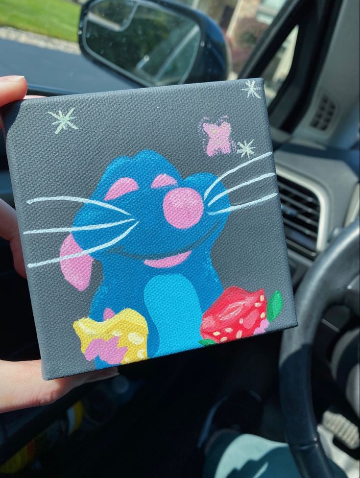 a person holding up a small square painting in their car's dash board holder