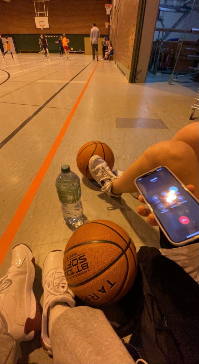 two basketballs are laying on the floor while someone is holding a cell phone and looking at them