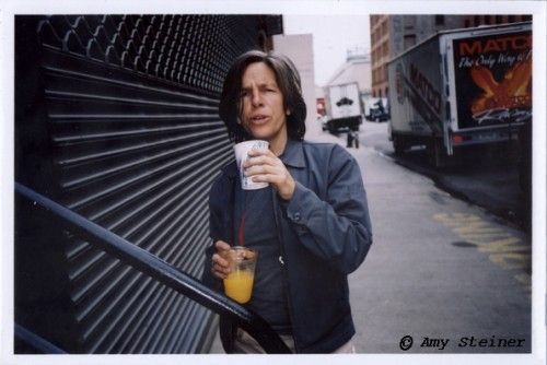 a woman holding a cup and standing next to a wall on the side of a street