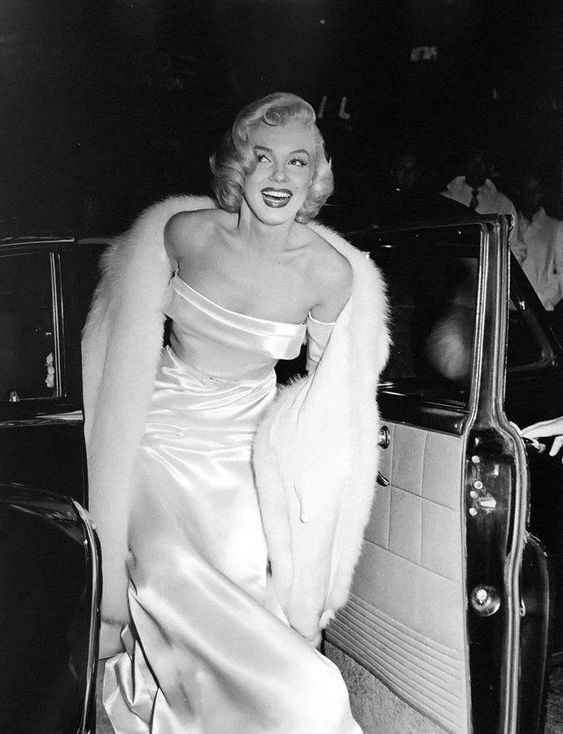 a woman in a white dress and fur stole around her shoulders, leaning against the door of a car