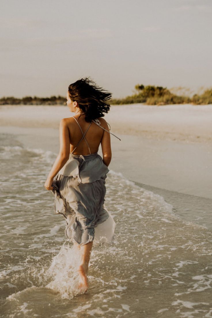 a woman is walking in the water at the beach with her dress flowing over her body