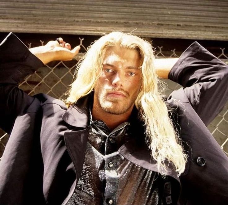 a man with long blonde hair standing next to a fence