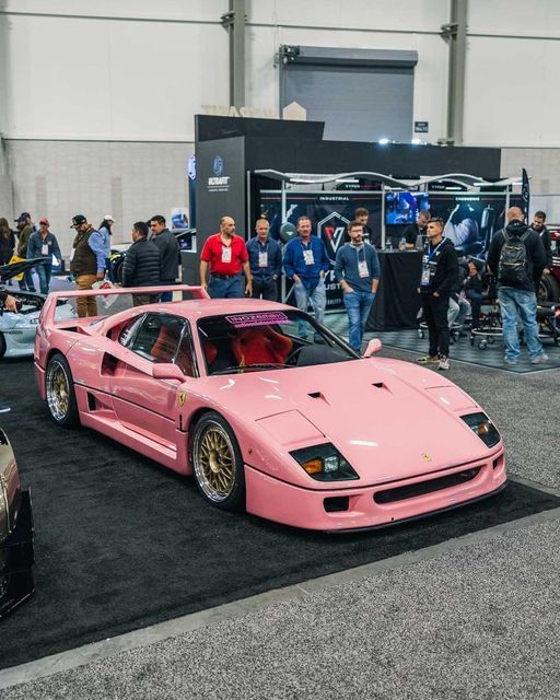 a pink sports car parked on top of a black carpet next to other cars and people