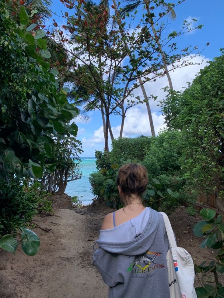 a woman walking down a dirt path next to the ocean with trees on either side