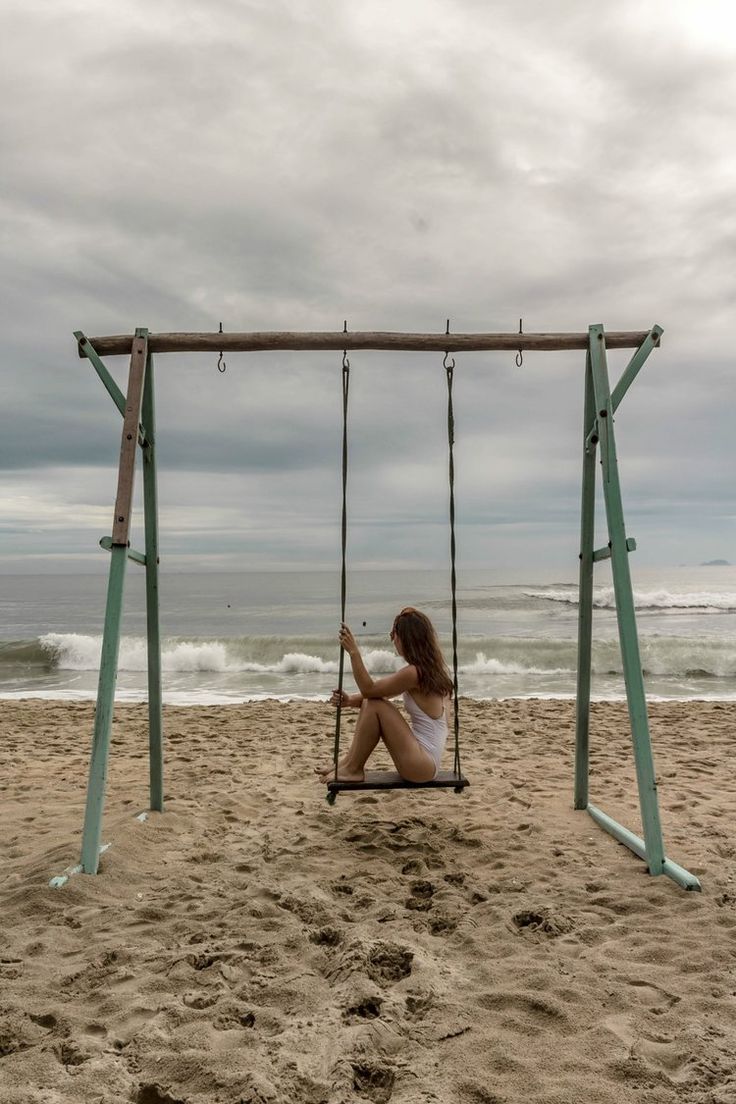 a woman is sitting on a swing at the beach with her feet in the sand