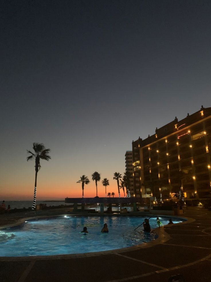 people are swimming in an outdoor pool at night with palm trees and hotel lights on the side