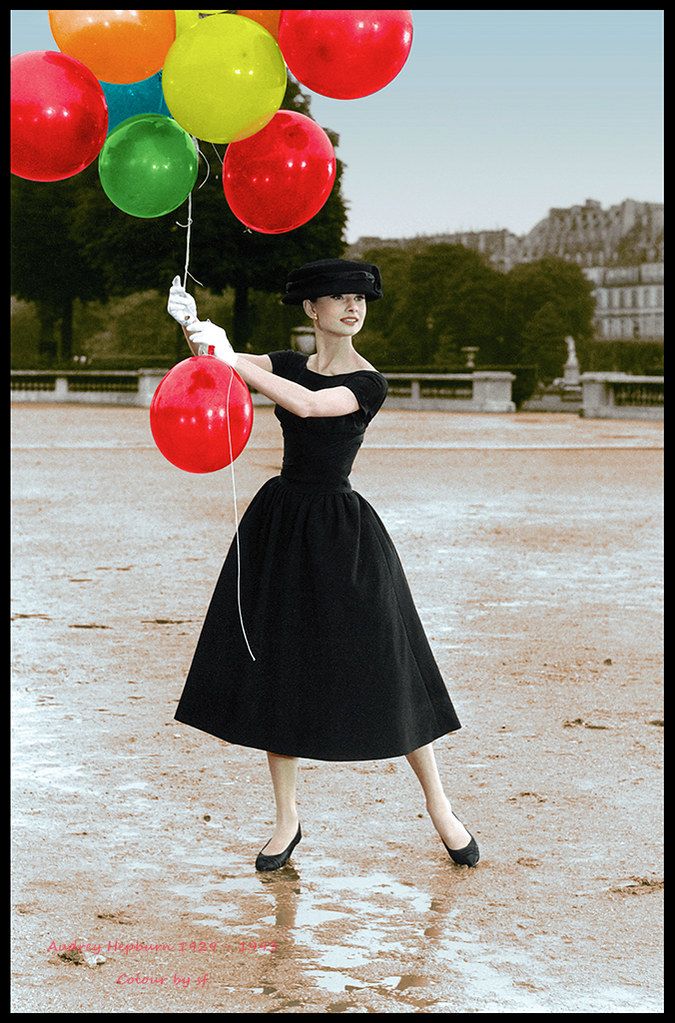 a woman in a black dress and hat holding bunches of balloons while standing on wet ground