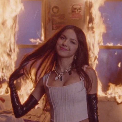 a woman in white dress and black leather gloves standing next to a wall with flames