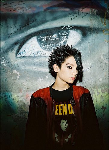 a young man with black hair standing in front of a graffiti covered wall