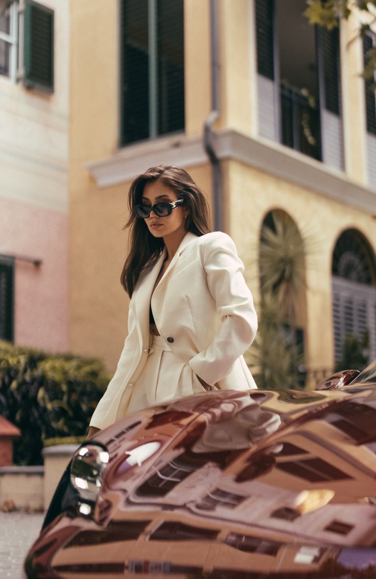 a woman in sunglasses is standing next to a car and looking at the camera while wearing a white coat