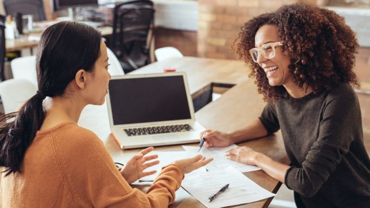 two women sitting at a table talking to each other and one is holding a pen