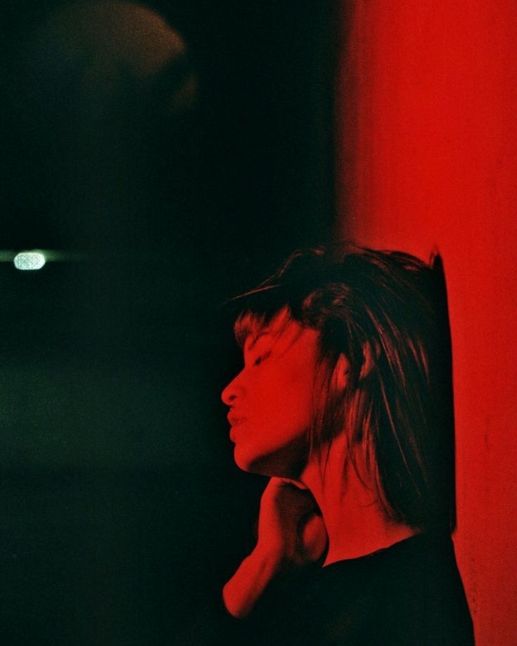 a woman standing in front of a red wall with her hand under her chin and looking off to the side