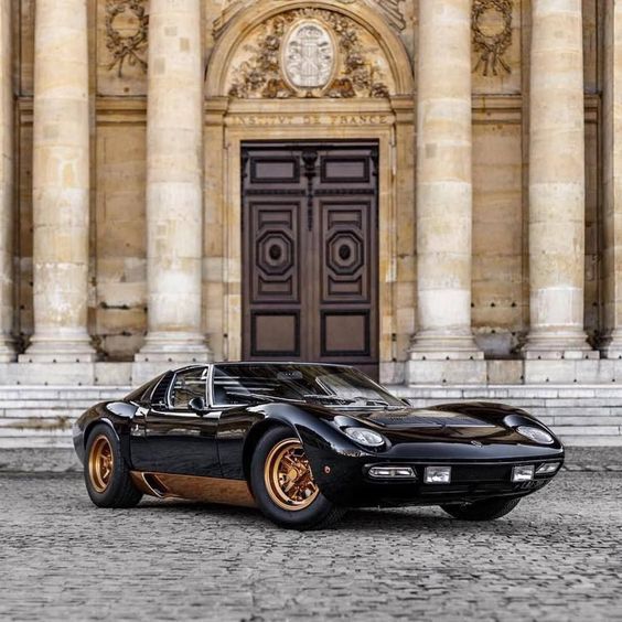 a black and gold sports car parked in front of a large building with columns on either side