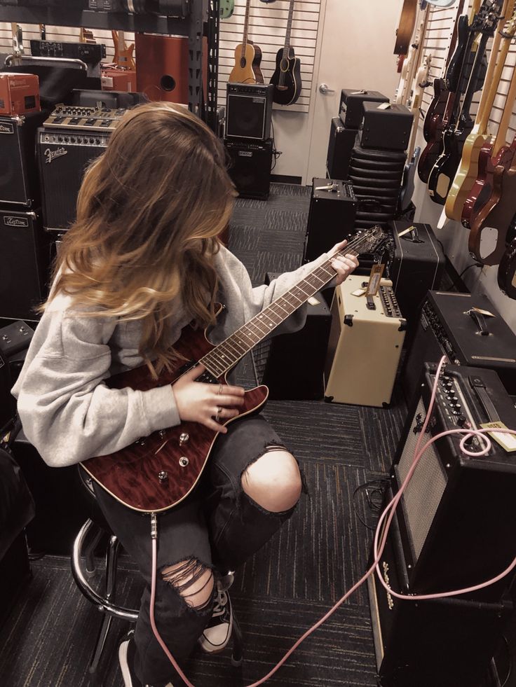 a woman sitting on the floor playing an electric guitar in a room full of guitars