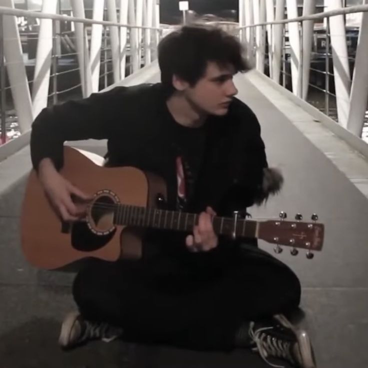 a young man sitting on the ground playing an acoustic guitar with his right hand and left arm