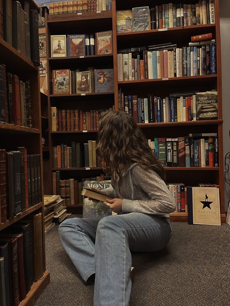 a woman sitting on the floor in front of a bookshelf reading a book