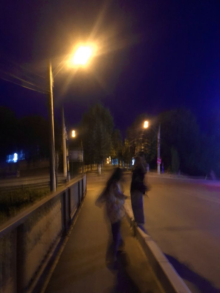 two people riding skateboards down a sidewalk at night time with street lights in the background
