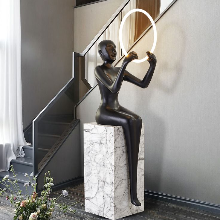 a statue holding a circular light in front of a stair case next to a window