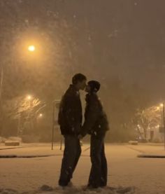 two people standing in the snow at night with street lights behind them and one person kissing the other