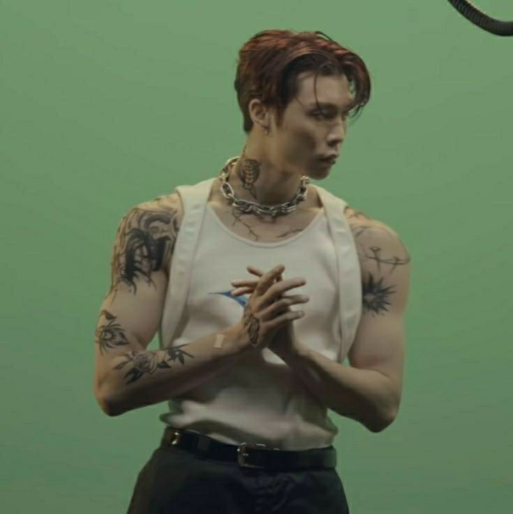 a man with tattoos and piercings standing in front of a green wall holding his hands together