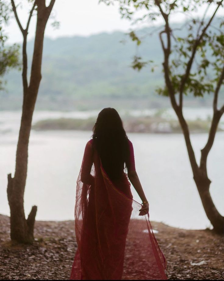 a woman in a pink sari is walking by some trees and the water behind her