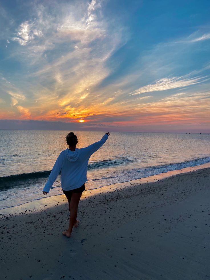 a woman walking on the beach at sunset with her arms in the air and one hand out
