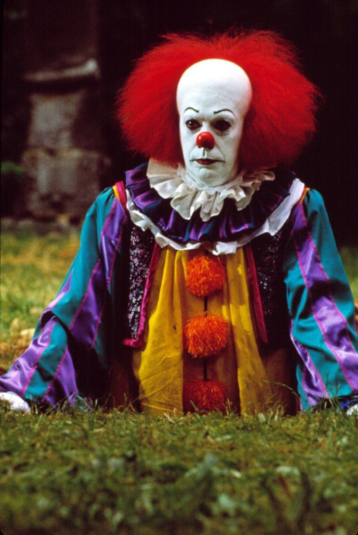 a creepy clown sitting in the grass with his head turned to look like he is wearing a red wig