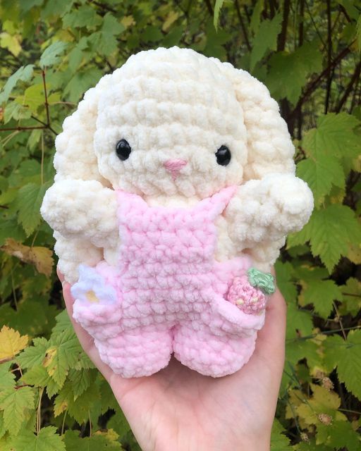 a hand holding a small crocheted white and pink stuffed animal in front of green leaves