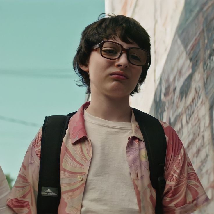 a young boy wearing glasses standing in front of a wall with graffiti writing on it