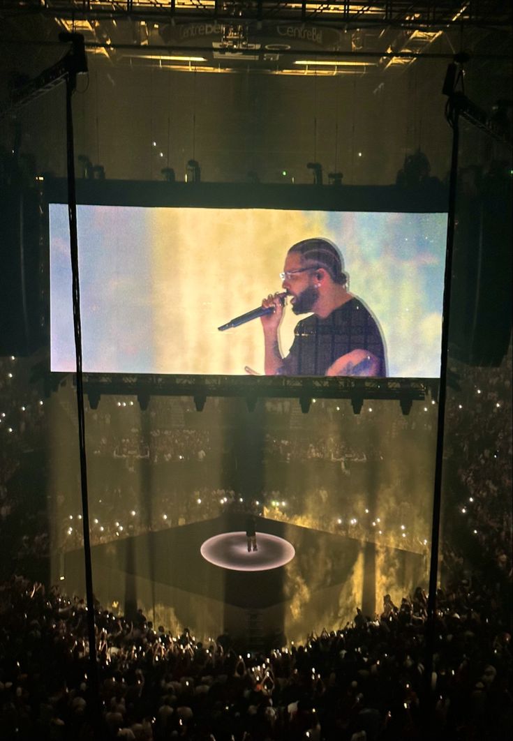 a person on a stage with a microphone in front of a large screen that shows a concert scene