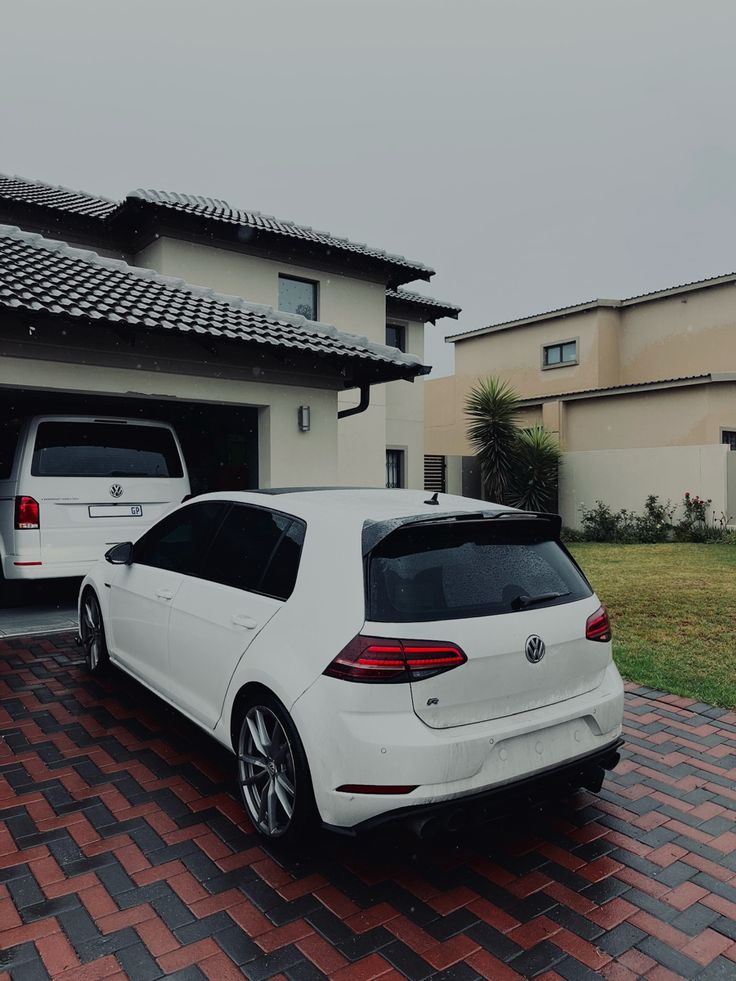 two white cars are parked in front of a house on a brick driveway next to a car garage