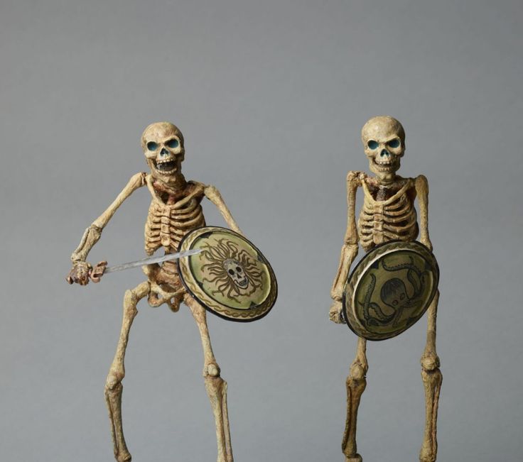 two skeleton figurines with shields and swords in front of grey background, one holding a shield