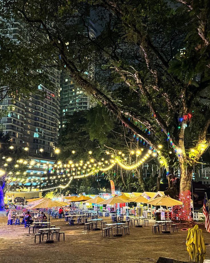 an outdoor event with tables and umbrellas lite up in the night, surrounded by tall buildings