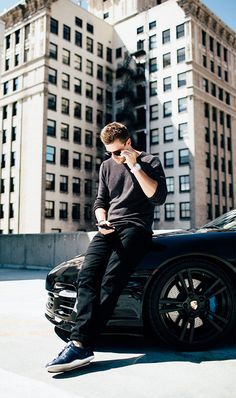 a man sitting on the hood of a black car talking on his cell phone in front of some tall buildings