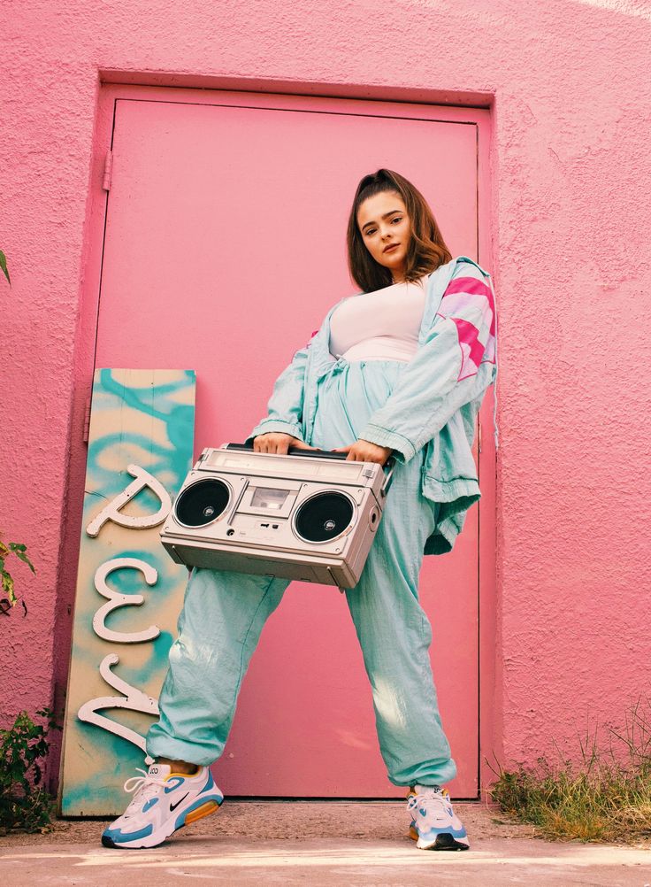 a woman standing in front of a pink wall holding an old school boombox and wearing blue sweatpants