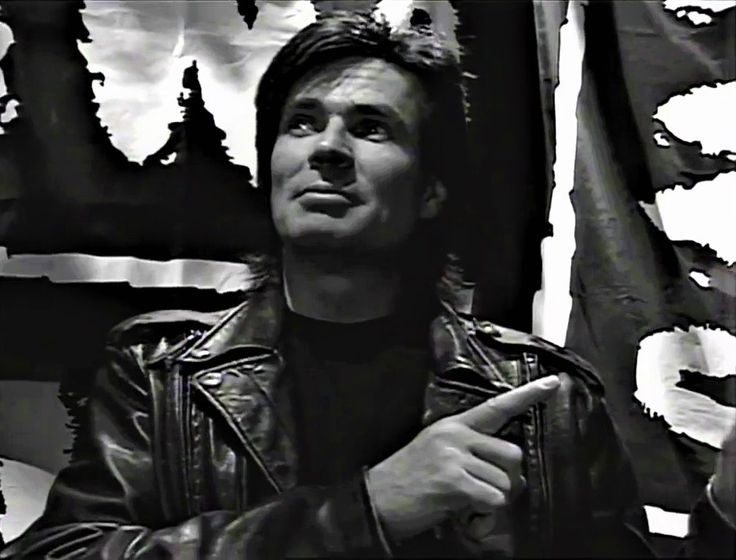 a black and white photo of a man in leather jacket pointing to the side with an american flag behind him