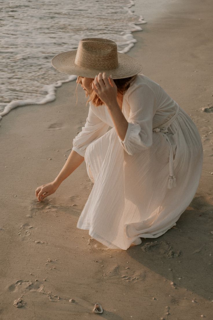 a woman in a white dress and hat crouches on the sand at the beach