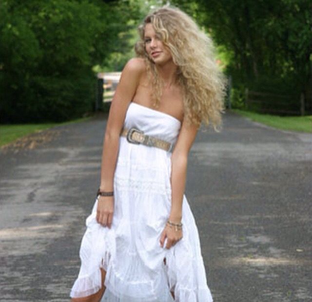 a woman in a white dress and cowboy boots posing for the camera on a country road