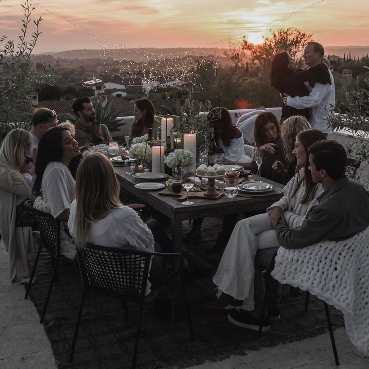 a group of people sitting around a table with food and drinks on it at sunset