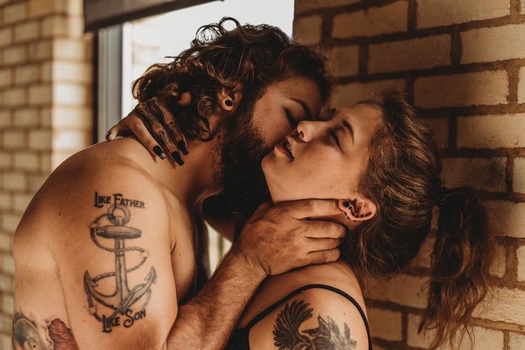 a man and woman with tattoos on their arms kissing each other in front of a brick wall