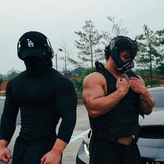 two men standing next to each other in front of a car wearing helmets and bodysuits