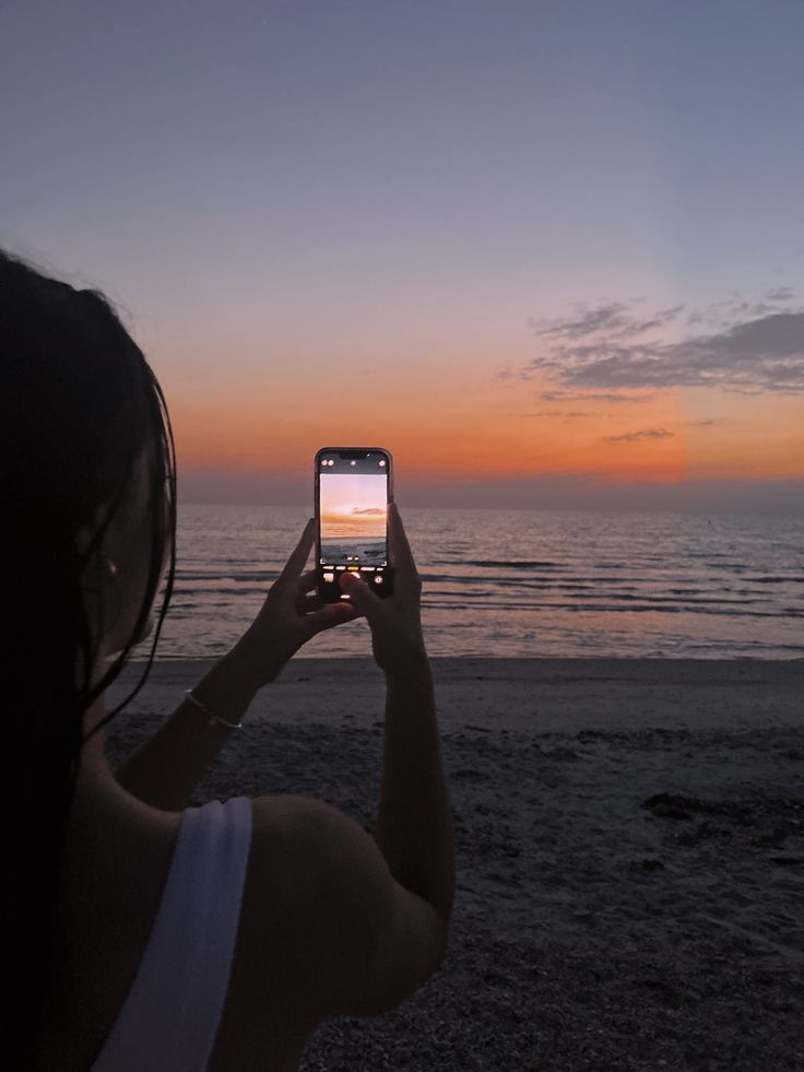 a woman taking a photo with her cell phone on the beach at sunset or dawn