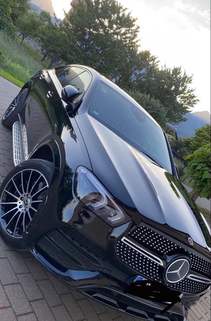 the front end of a black car parked on a brick road with trees in the background